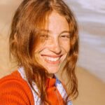 Close-up portrait of a smiling woman with freckles and red hair at the beach during the day.